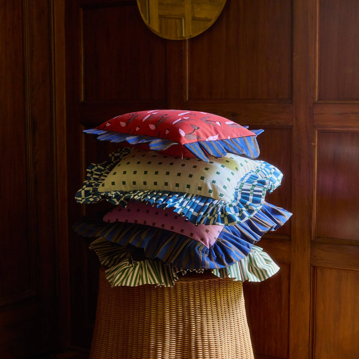 Pink Checks Ruffle Pillow in a stack of Piecework pillows on a wicker table