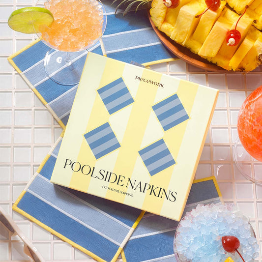 A box of Piecework Poolside Napkins displayed over light and medium blue embroidered linen cocktail napkins with wide cabana stripes and yellow trim, surrounded by frozen cocktails and sliced pineapple on white pool tiles.