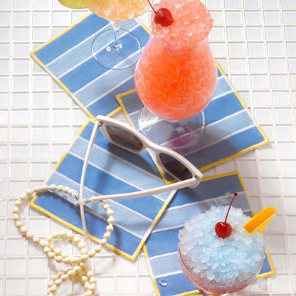Light and medium blue embroidered linen cocktail napkins with wide cabana stripes and yellow trim, styled on clear mosaic tiles with frozen cocktails, white sunglasses, and a pearl necklace.