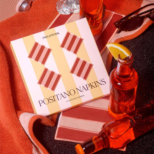 A boxed set of Positano Napkins by Piecework styled on a dark surface with orange towels, a glass bottle of red drink, sunglasses, and an orange garnish. The napkin box has a cream and yellow striped background with diagonal salmon and orange striped squares.

