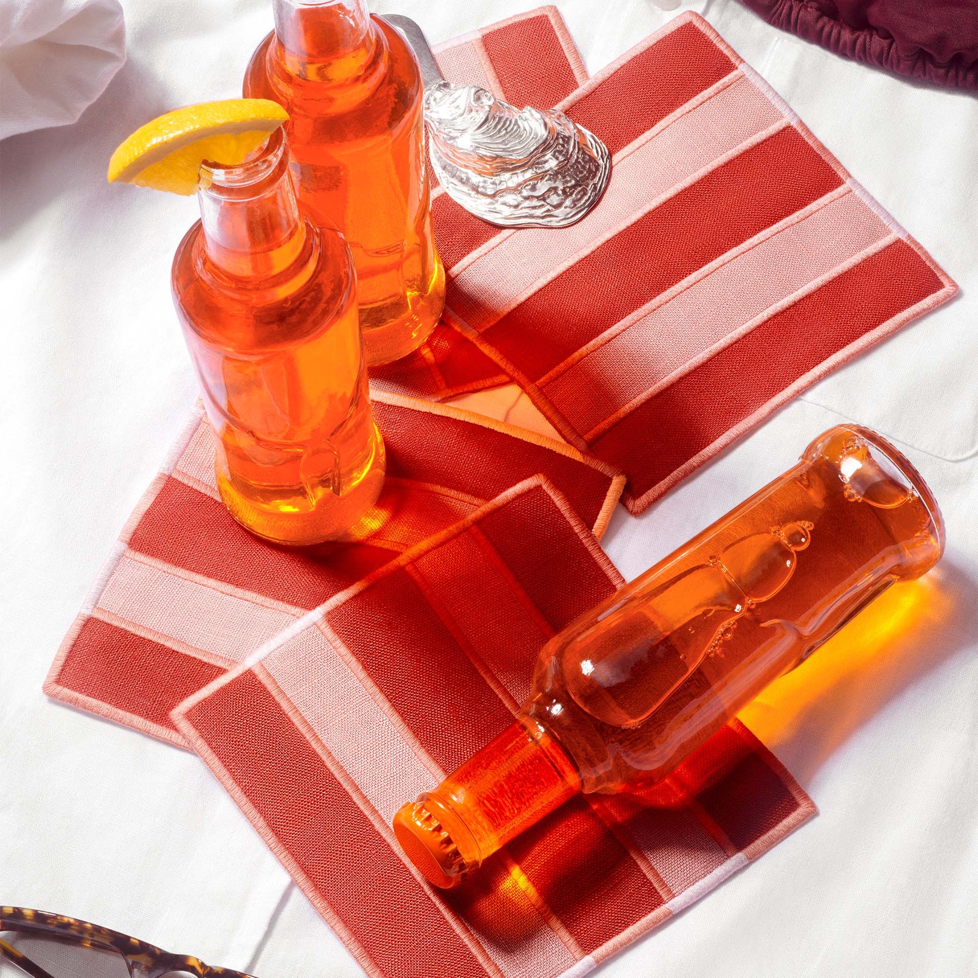 Deep terracotta and salmon striped woven linen embroidered cocktail napkins styled with two clear bottles filled with orange liquid, a orange slice on a white fabric surface.