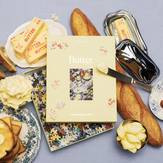 Butter-themed puzzle box with silver foil lettering along with sticks of butter, a loaf of bread, and other bread and butter items on a white tablecloth background.