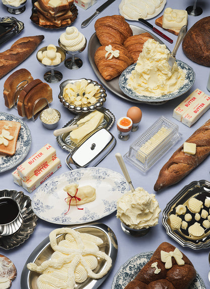 A spread of butter, assorted pastries, and bread on a table along with silver dishes and white ceramic platters with blue details.