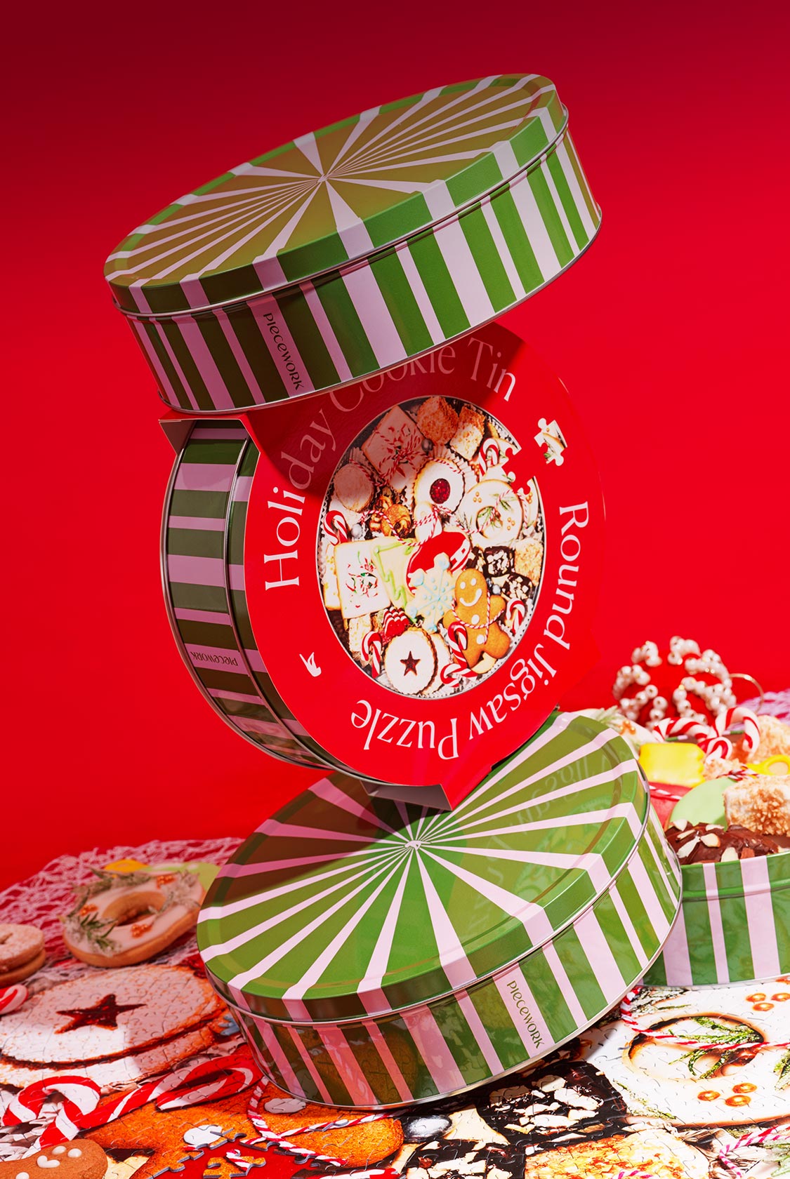 Piecework Puzzles' new 750-piece round jigsaw puzzle (Holiday Cookie Tin) stacked in a vibrant red scene on top of the assembled puzzle.