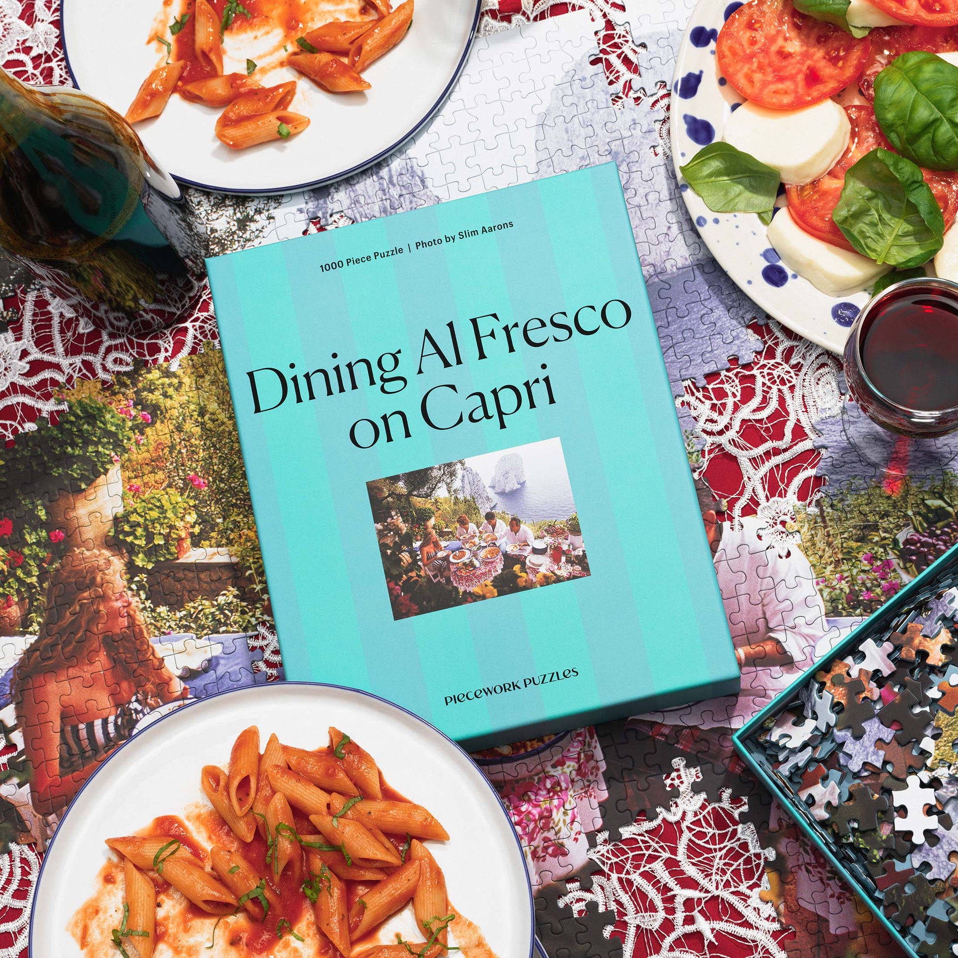 Top-down view of the 'Dining al Fresco on Capri' 1000-piece puzzle box, laid flat on a table featuring a partially assembled puzzle, an open box of puzzle pieces, and a fresh Caprese salad, capturing the Mediterranean charm of Capri.