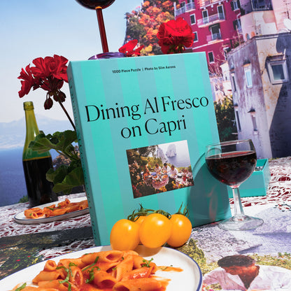 Blue-green striped puzzle box for 'Dining al Fresco on Capri' 1000-piece puzzle, featuring a Mediterranean tablescape scene with vibrant colors and sun-soaked elegance, evoking the effortless glamour of Capri.
