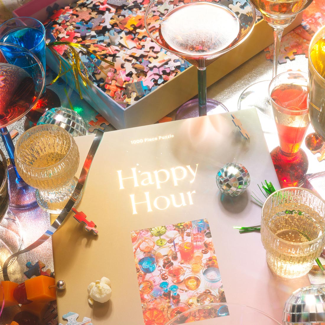 A festive tabletop scene featuring the “Happy Hour” 1000-piece jigsaw puzzle by Piecework. The puzzle box sits surrounded by colorful cocktail glasses, disco balls, puzzle pieces, and glimmering reflections. Bright light bounces across the metallic surface, creating a playful, celebratory mood reminiscent of a vibrant party setup.