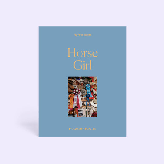A single jigsaw puzzle box standing upright on a pale lavender background. The box is dusty blue with the title 'Horse Girl' in large gold text and a small central collage photo of horse girl memorabilia.