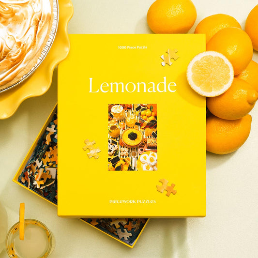 Open puzzle box for Lemonade, a 1000-piece puzzle, shown on a white surface surrounded by lemons and desserts.