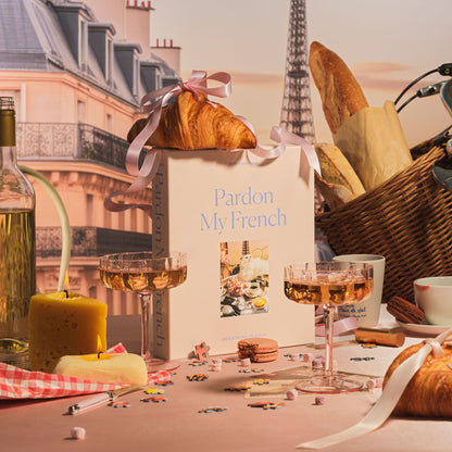 A Parisian-inspired gift selection featuring Piecework's 'Pardon My French' puzzle, buttery croissants, champagne coupes, artisanal cheese, and classic French café elements—perfect for lovers of French culture and elegant gifting.