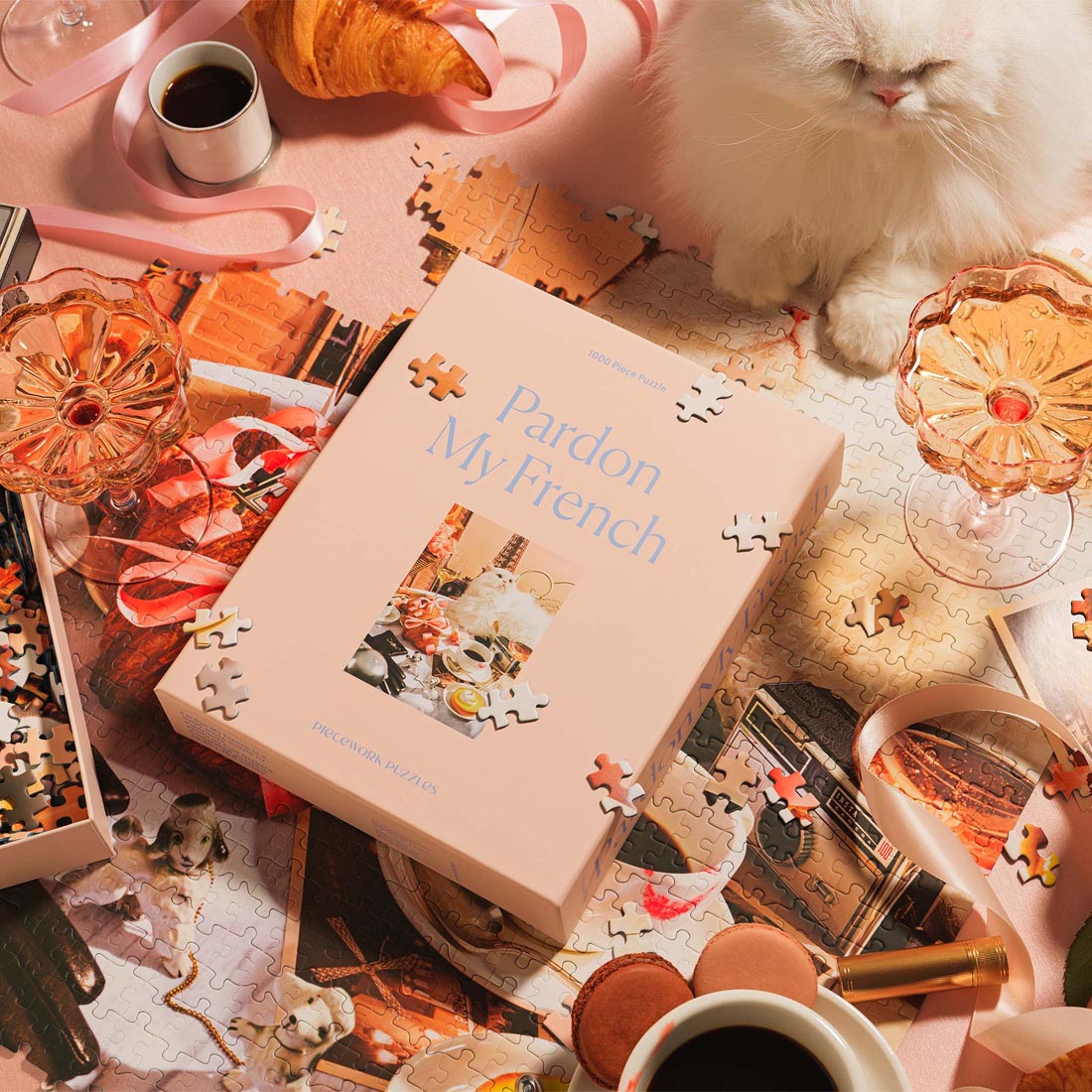 A chic tabletop scene featuring the “Pardon My French” 1000-piece jigsaw puzzle by Piecework Puzzles. The soft pink puzzle box is surrounded by scattered puzzle pieces, croissants, macarons, coffee cups, pink glassware, and satin ribbon. A fluffy white cat sits nearby, completing the elegant Parisian-inspired vignette.