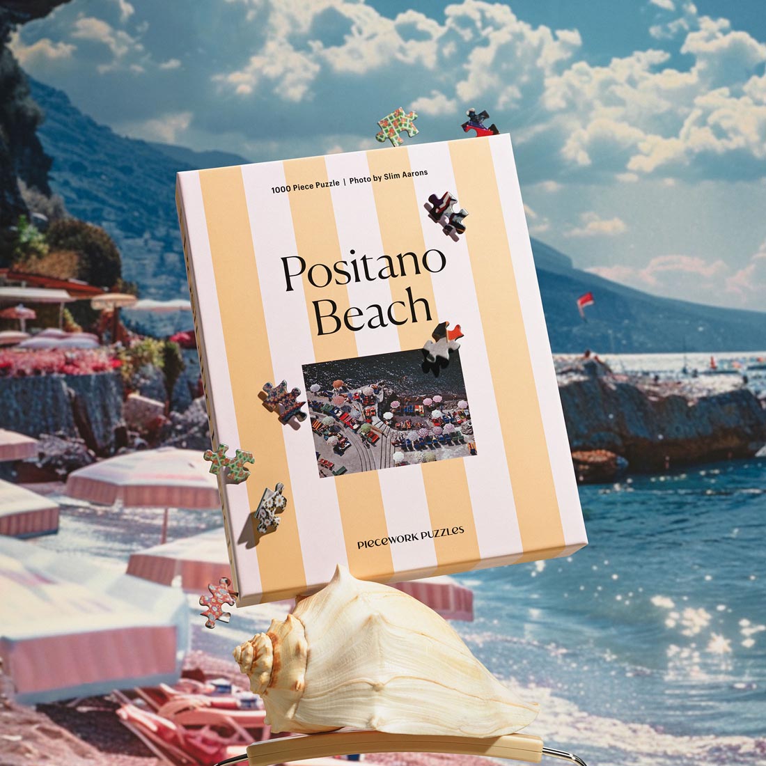 A 1000-piece puzzle box featuring an iconic Slim Aarons photograph of sunbathers and parasols on Positano Beach, with the box placed on a real beach setting.
