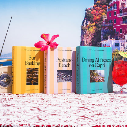 View into a Mediterranean table setting featuring the 'Dining al Fresco on Capri,' 'Sun Basking,' and 'Positano Beach' 1000-piece puzzle boxes, showcasing the distinctive cabana stripe packaging of the Piecework's Slim Aarons puzzle collection amidst a relaxed, sunlit Mediterranean scene.