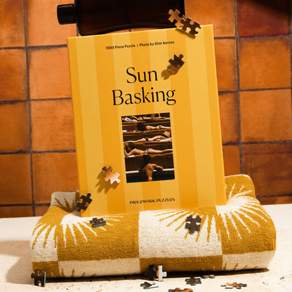 A puzzle box with the title 'Sun Basking' displayed on a surface, featuring a image of people sunbathing. The box on top of a folded beach towel with a few puzzle pieces visible.
