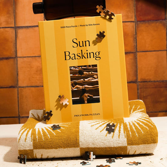 A puzzle box with the title 'Sun Basking' displayed on a surface, featuring a image of people sunbathing. The box on top of a folded beach towel with a few puzzle pieces visible.