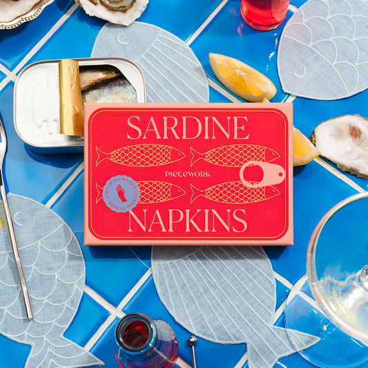 Packaging for Sardine Napkins on a blue tile counter top. The box for the napkins are surrounded by fish-shaped linen napkins and seafood, lemons and drinkware.
