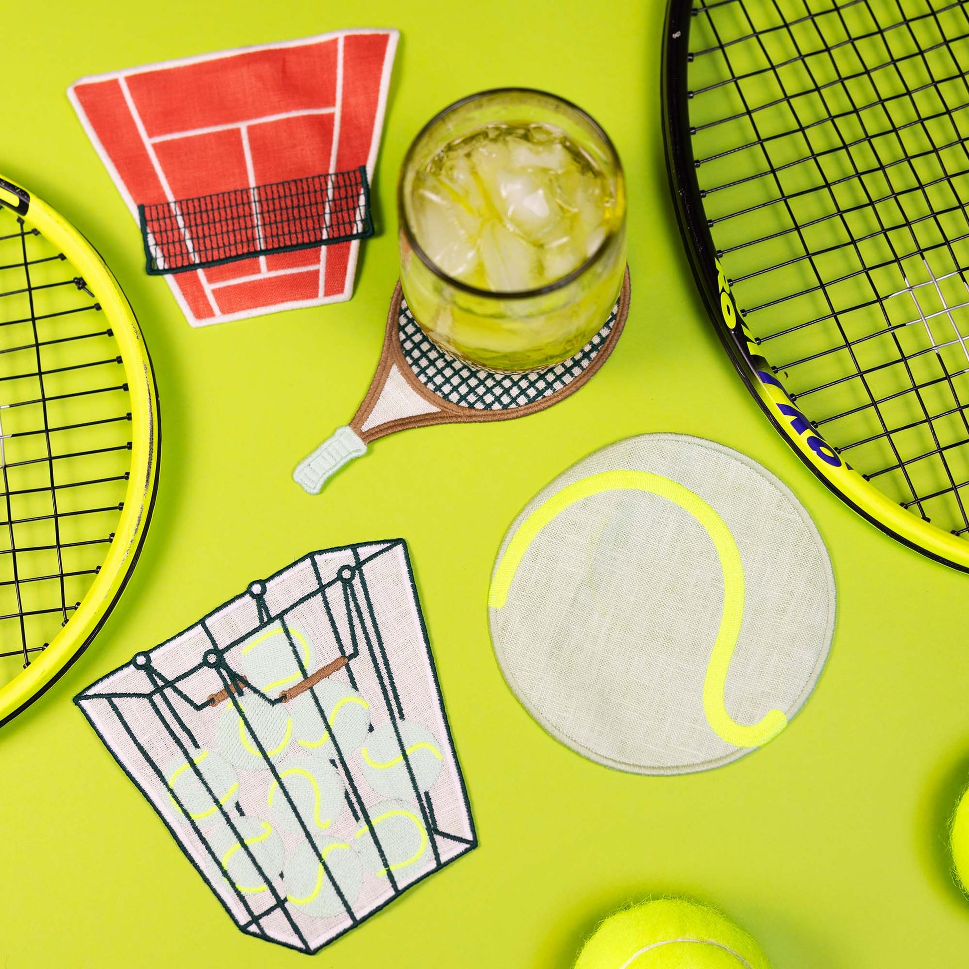 Tennis-themed cocktail napkins paired with rackets, a ball, and a glass of lemonade on a neon green surface.