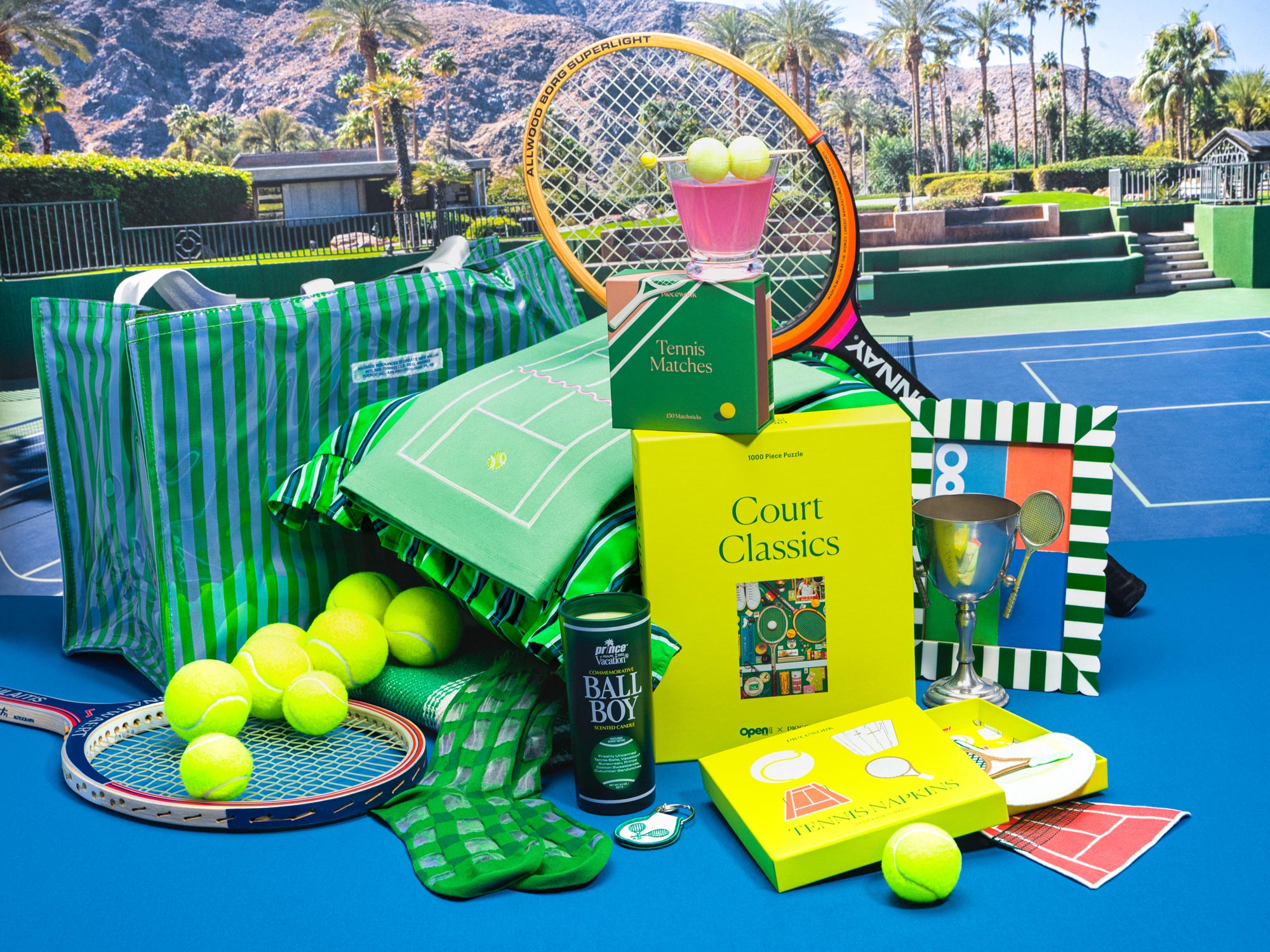 Tennis-themed products including rackets, balls, candles, puzzles, matches, and cocktail napkins on a blue court surface.