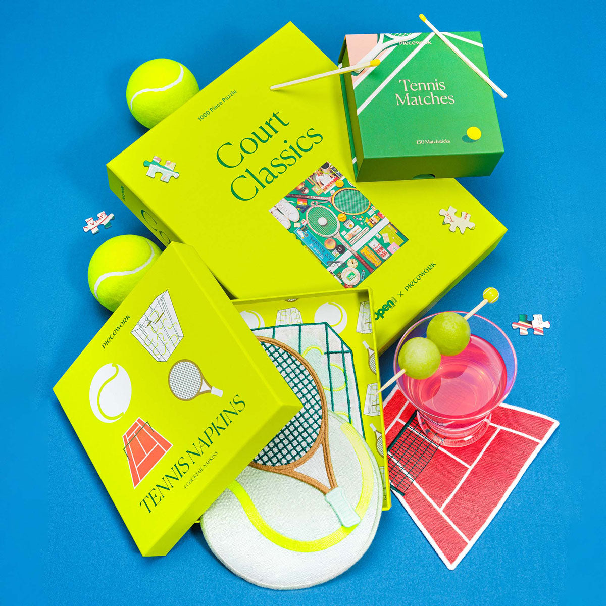 The Tennis Set featuring the Court Classics puzzle, Tennis Matches, and embroidered Tennis Cocktail Napkins styled with tennis balls, puzzle pieces, and a pink cocktail on a bright blue background.