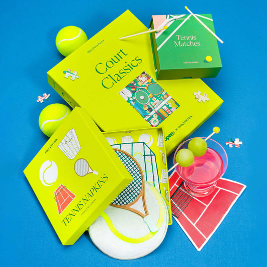 The Tennis Set featuring the Court Classics puzzle, Tennis Matches, and embroidered Tennis Cocktail Napkins styled with tennis balls, puzzle pieces, and a pink cocktail on a bright blue background.