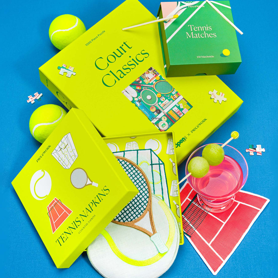A zoomed in view of The Tennis Set featuring the Court Classics puzzle, Tennis Matches, and embroidered Tennis Cocktail Napkins styled with tennis balls, puzzle pieces, and a pink cocktail on a bright blue background.