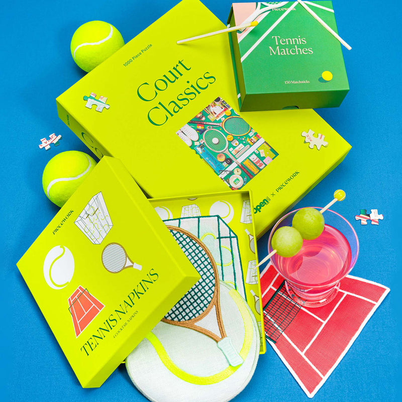 A zoomed in view of The Tennis Set featuring the Court Classics puzzle, Tennis Matches, and embroidered Tennis Cocktail Napkins styled with tennis balls, puzzle pieces, and a pink cocktail on a bright blue background.