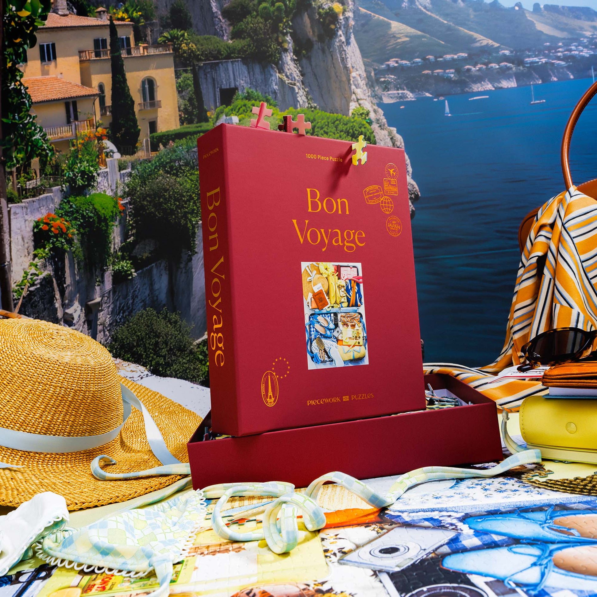 Bon Voyage puzzle box displayed in a travel-inspired scene with a scenic coastal cliffs backdrop, straw hat, striped scarf, sunglasses, and assembled puzzle underneath.