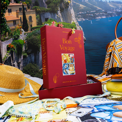 Bon Voyage puzzle box displayed in a travel-inspired scene with a scenic coastal cliffs backdrop, straw hat, striped scarf, sunglasses, and assembled puzzle underneath.