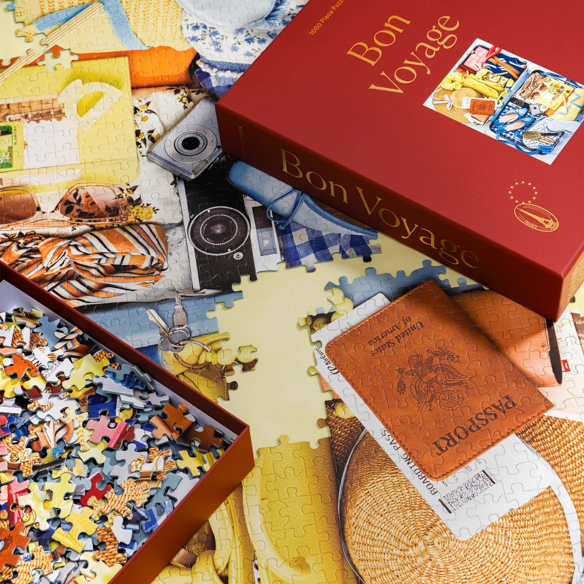 Bon Voyage puzzle in progress with the burgundy red puzzle box and loose pieces styled across the in progress puzzle, puzzle art is filled with a passport, camera, and travel accessories.