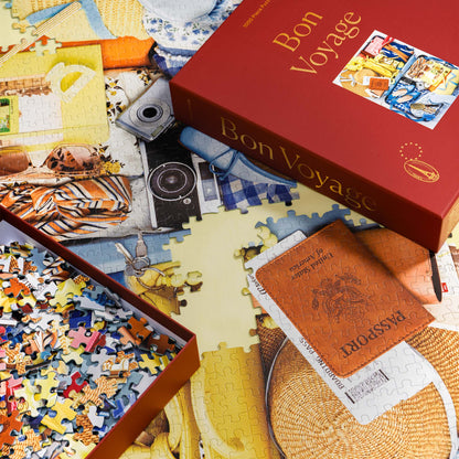 Bon Voyage puzzle in progress with the burgundy red puzzle box and loose pieces styled across the in progress puzzle, puzzle art is filled with a passport, camera, and travel accessories.