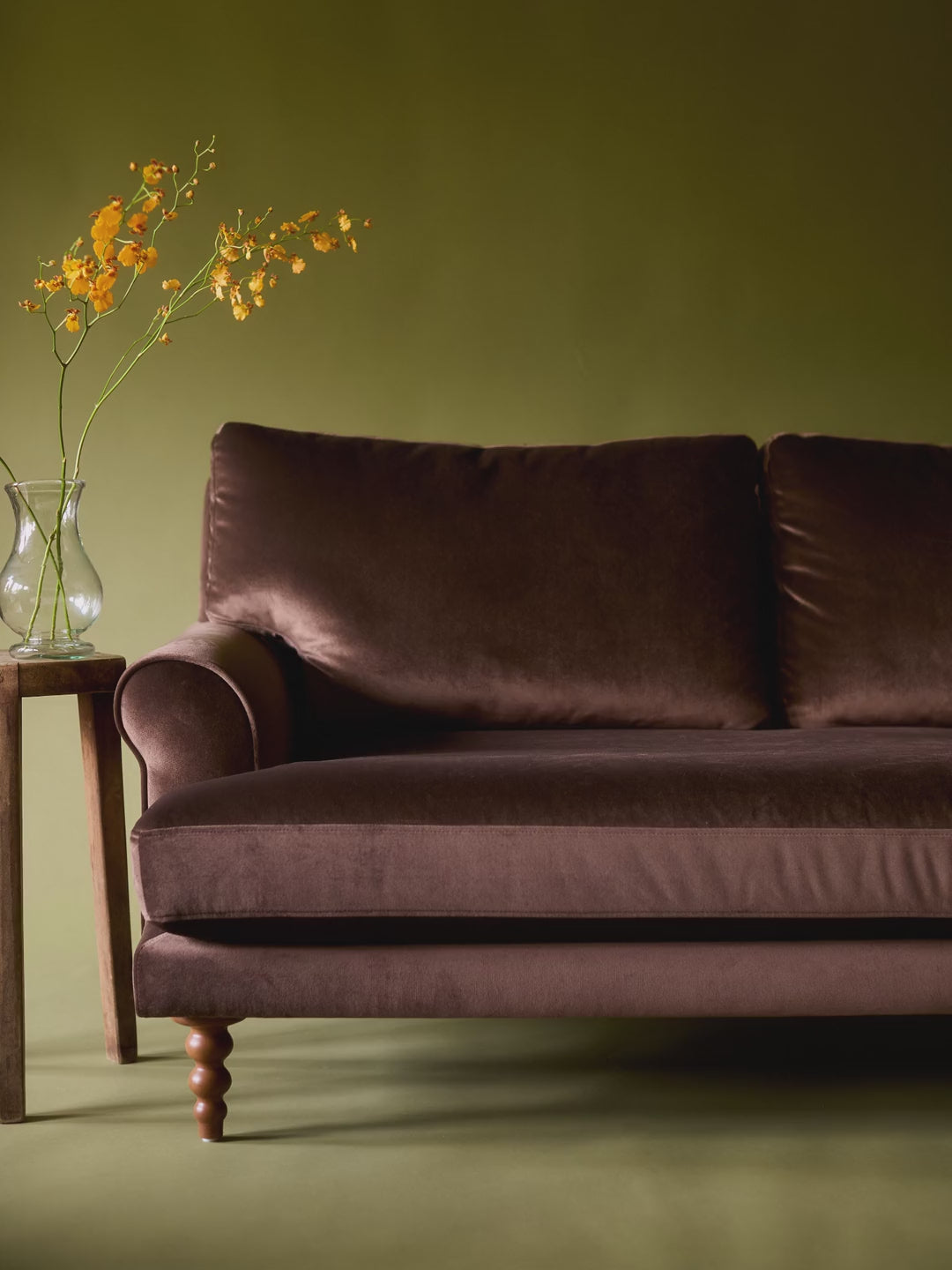 Piecework petal and ruffle pillows piling up one by one on a brown velvet sofa beside a vase of yellow orchids