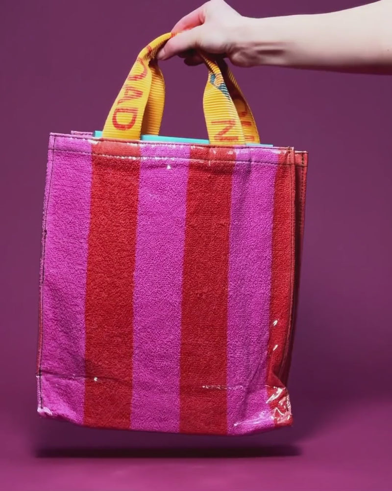 A hand holding the yellow woven handles of a magenta and red striped tote bag with a clear vinyl exterior, against a magenta background.