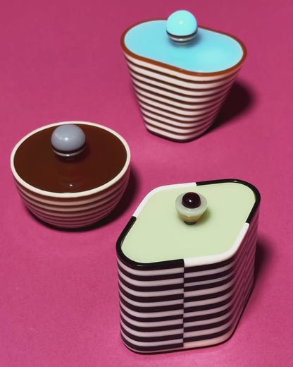 A hand reaching into a small striped bowl of candy beside a black-and-white striped rectangular box filled with a rainbow gummy candy, on a magenta surface.