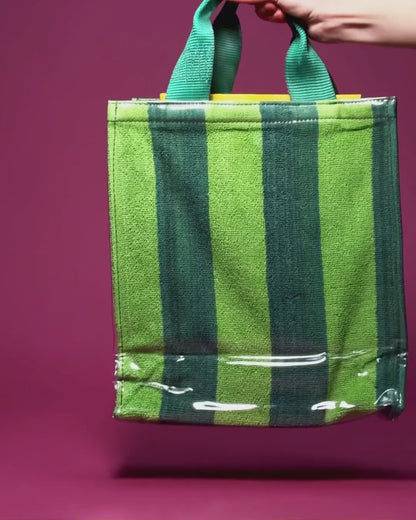 A hand holding the green woven handles of a green and dark green striped tote bag with a clear vinyl exterior, against a magenta background.