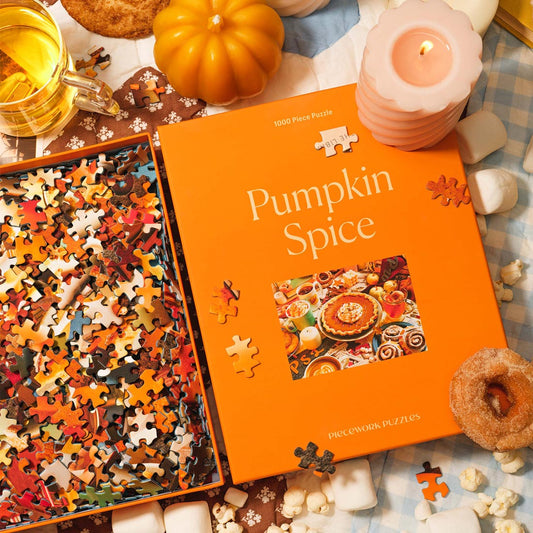 A cozy autumn-themed scene featuring the “Pumpkin Spice” 1000-piece jigsaw puzzle by Piecework Puzzles. The bright orange box sits open with colorful puzzle pieces spilling out, surrounded by a lit candle, mini pumpkin, mug of tea, donut, popcorn, and marshmallows. Warm tones and fall textures evoke a festive, seasonal mood.