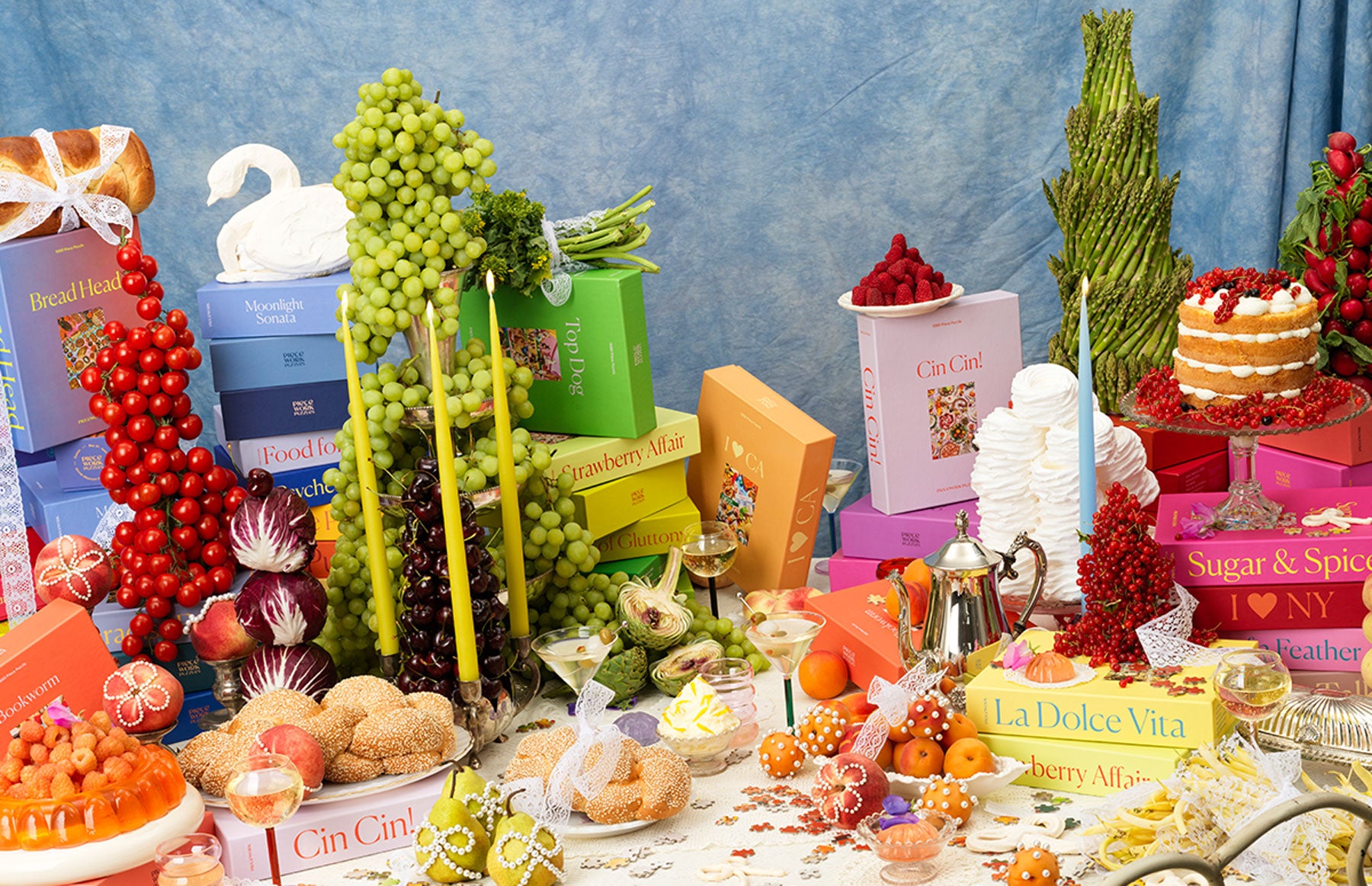 A colorful Piecework puzzle feast tablescape with stacked puzzle boxes, fruit towers, candles, cakes, breads, asparagus, radishes, whipped cream, and vintage serving pieces arranged against a blue backdrop.