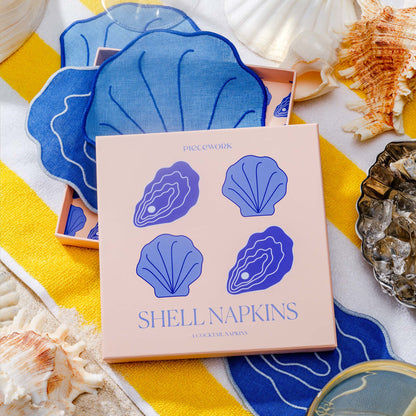 Piecework Shell Napkins gift box with blue clam and oyster napkins styled on beach towel with seashells.