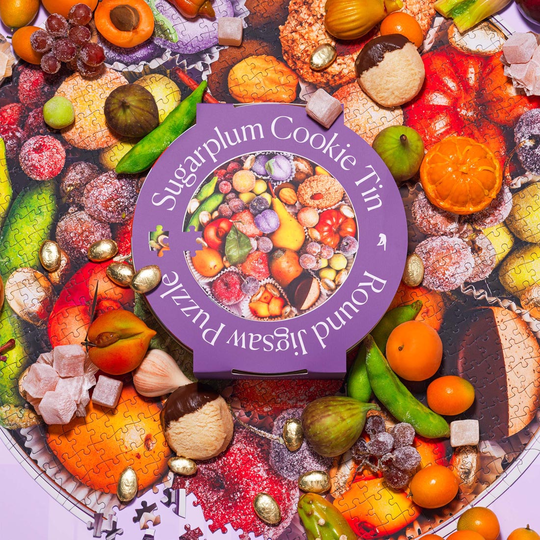 A vibrant, round jigsaw puzzle titled “Sugarplum Cookie Tin” by Piecework Puzzles. The limited-edition puzzle tin sits on top of the assembled puzzle, surrounded by colorful sweets, figs, citrus fruits, candied treats, and gold-wrapped chocolates. The scene evokes a festive, confectionary display full of texture and holiday cheer.